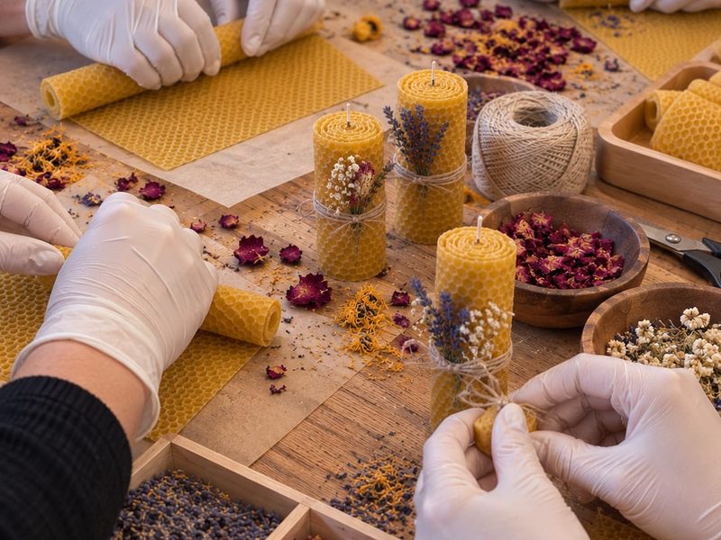 Gloved hands rolling beeswax candle sheets at a workshop table scattered with dried flowers, lavender and twine