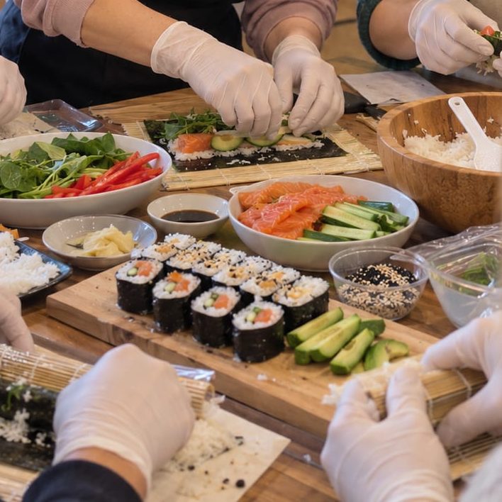 Gloved hands rolling sushi on bamboo mats beside bowls of rice, vegetables and prepared ingredients